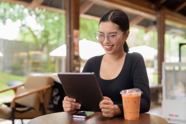 Tablet bilgisayarla çalışan ve kahve dükkanında buzlu Tayland çayı içen güzel bir Asyalı kadın.