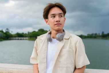 Kulaklıklı Asyalı genç turist göl kenarındaki parkta dinleniyor.