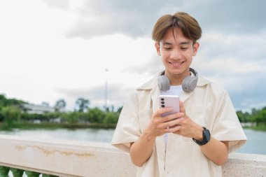 Kulaklıklı Asyalı genç turist, göl kenarındaki parkta telefonla dinleniyor.