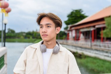 Kulaklıklı Asyalı genç turist göl kenarındaki parkta dinleniyor.