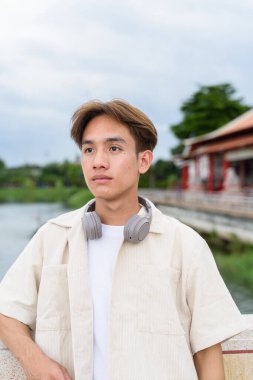 Kulaklıklı Asyalı genç turist göl kenarındaki parkta dinleniyor.