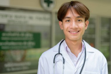 Genç Asyalı sağlık çalışanı kliniğin önünde laboratuvar önlüğü giyiyor.