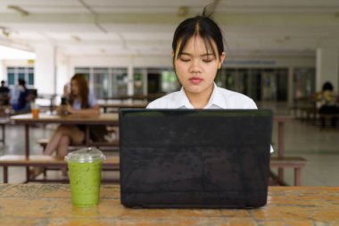 Üniversite üniformalı Taylandlı genç kız kampüsün kahve dükkanında tek başına oturuyor. Eğitimsel bir ortamda gerçek öğrenci hayatını, güzelliğini ve gençliğini gösteren rahat bir yaşam tarzı portresi..