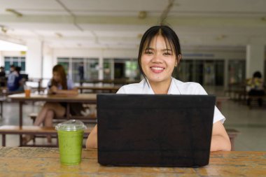 Üniversite üniformalı Taylandlı genç kız kampüsün kahve dükkanında tek başına oturuyor. Eğitimsel bir ortamda gerçek öğrenci hayatını, güzelliğini ve gençliğini gösteren rahat bir yaşam tarzı portresi..