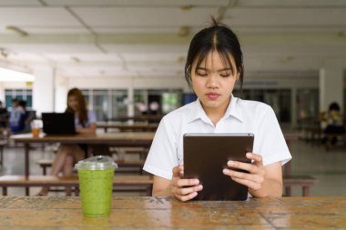 Üniversite üniformalı Taylandlı genç kız kampüsün kahve dükkanında tek başına oturuyor. Eğitimsel bir ortamda gerçek öğrenci hayatını, güzelliğini ve gençliğini gösteren rahat bir yaşam tarzı portresi..