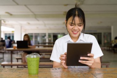Üniversite üniformalı Taylandlı genç kız kampüsün kahve dükkanında tek başına oturuyor. Eğitimsel bir ortamda gerçek öğrenci hayatını, güzelliğini ve gençliğini gösteren rahat bir yaşam tarzı portresi..