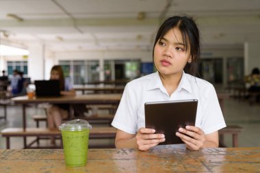 Üniversite üniformalı Taylandlı genç kız kampüsün kahve dükkanında tek başına oturuyor. Eğitimsel bir ortamda gerçek öğrenci hayatını, güzelliğini ve gençliğini gösteren rahat bir yaşam tarzı portresi..