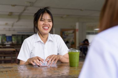 Üniversite öğrencisi üniformalı iki güzel Taylandlı kampüsün açık hava kahve dükkanında birlikte oturuyorlar. Rahatlamış, samimi bir an. Arkadaşlık ya da çift etkileşimi gösteriyor..
