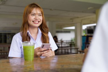 Üniversite öğrencisi üniformalı iki güzel Taylandlı kampüsün açık hava kahve dükkanında birlikte oturuyorlar. Rahatlamış, samimi bir an. Arkadaşlık ya da çift etkileşimi gösteriyor..