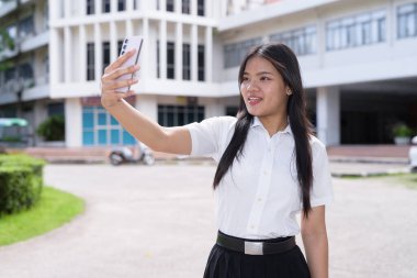 Güzel Tayland üniversitesi öğrencisi kampüste tek başına duruyor ve resmi akademik üniforma giyiyor. Gençlik, bağımsızlık ve akademik hırs odaklı otantik, modern eğitim teması.