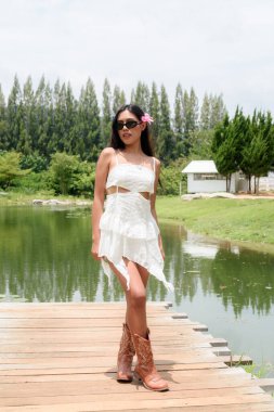 Beyaz elbiseli ve botlu genç Asyalı bir kadının tam vücut portresi göl manzaralı. Rahat turist havası, özgürlük ve kadınsı seyahat tarzıyla doğada şık bir yaz tatili konsepti..