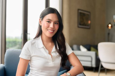 Beyaz polo tişörtlü güzel bir Asyalı kadın otel veya restoran salonunda poz veriyor. Modern iç mekan, tarz ve doğal ifade ile rahat seyahat tarzı portresi.