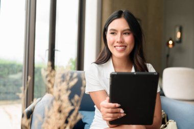 Otel ya da kafe salonunda otururken tablet kullanan genç ve güzel bir Asyalı turist. Modern iç mekan ve kendine güvenen doğal ifade ile günlük, şık dijital yaşam tarzı.