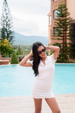 Beyaz polo tişörtlü genç Asyalı kadın güneşli doğa ortamında havuz kenarında poz veriyor. Yeşil arka plan, doğal ışık ve sakin turist ifadesine sahip gündelik yaz tatili atmosferi.