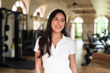 Beyaz polo tişörtlü güzel bir Asyalı kadın spor salonunda poz veriyor. Arka planda modern ekipmanlar olan şık fitness yaşam tarzı konsepti. Rahat ve sportif, egzersiz yapmayan moda portresi..