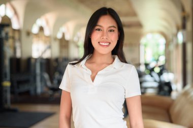 Beyaz polo tişörtlü güzel bir Asyalı kadın spor salonunda poz veriyor. Arka planda modern ekipmanlar olan şık fitness yaşam tarzı konsepti. Rahat ve sportif, egzersiz yapmayan moda portresi..