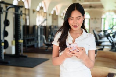 Beyaz polo tişörtlü güzel bir Asyalı kadın spor salonunda telefonla poz veriyor. Arka planda modern ekipmanlar olan şık fitness yaşam tarzı konsepti. Rahat ve sportif, egzersiz yapmayan moda portresi..