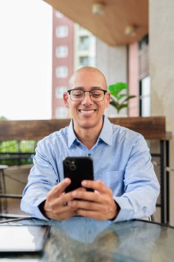 Gözlüklü, mavi tişörtlü, güpegündüz dışarıda oturan kendine güvenen kel bir adam. Yaşam tarzı ve iş teması.