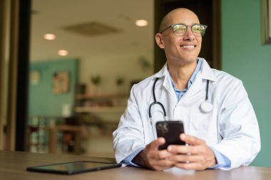 Gözlüklü ve laboratuvar önlüklü kel erkek doktor hastane veya klinik kütüphanesinin bekleme salonunda oturuyor. Sakin ve profesyonel ruh hali.