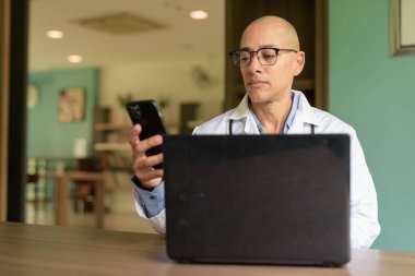 Gözlüklü ve laboratuvar önlüklü kel erkek doktor hastane veya klinik kütüphanesinin bekleme salonunda oturuyor. Sakin ve profesyonel ruh hali.