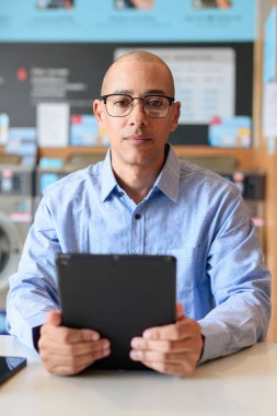 Mavi gömlekli ve gözlüklü kel adam modern çamaşırhanede otururken tablet bilgisayar kullanıyor. Dijital yaşam tarzı ve gerçek iş konsepti.