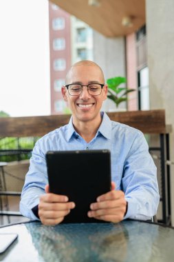 Mavi gömlekli ve gözlüklü kel adam gün içinde dışarıda otururken dijital tablet kullanıyor. Mobil teknoloji ve modern çalışma tarzı.