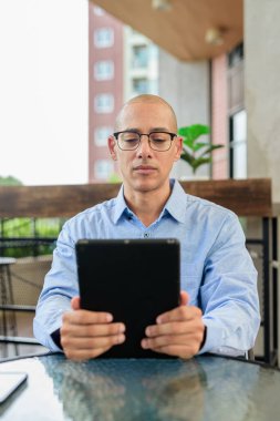 Mavi gömlekli ve gözlüklü kel adam gün içinde dışarıda otururken dijital tablet kullanıyor. Mobil teknoloji ve modern çalışma tarzı.