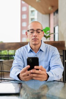Gözlüklü, mavi tişörtlü, güpegündüz dışarıda oturan kendine güvenen kel bir adam. Yaşam tarzı ve iş teması.