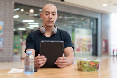Normal ortamlarda dijital tablet bilgisayar ve salata kullanan sağlıklı yetişkin bir adam. Gıda mahkemelerinde teknoloji, beslenme ve modern yaşam tarzı arasındaki dengeyi vurgular.