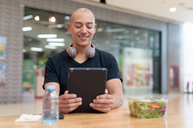 Normal ortamlarda dijital tablet bilgisayar ve salata kullanan sağlıklı yetişkin bir adam. Gıda mahkemelerinde teknoloji, beslenme ve modern yaşam tarzı arasındaki dengeyi vurgular.