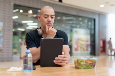 Normal ortamlarda dijital tablet bilgisayar ve salata kullanan sağlıklı yetişkin bir adam. Gıda mahkemelerinde teknoloji, beslenme ve modern yaşam tarzı arasındaki dengeyi vurgular.