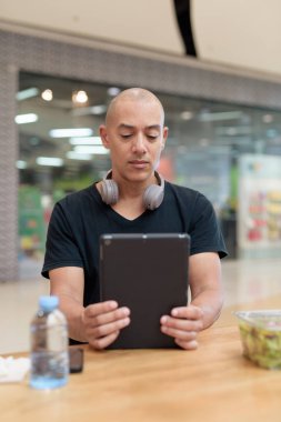 Normal ortamlarda dijital tablet bilgisayar ve salata kullanan sağlıklı yetişkin bir adam. Gıda mahkemelerinde teknoloji, beslenme ve modern yaşam tarzı arasındaki dengeyi vurgular.