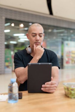 Normal ortamlarda dijital tablet bilgisayar ve salata kullanan sağlıklı yetişkin bir adam. Gıda mahkemelerinde teknoloji, beslenme ve modern yaşam tarzı arasındaki dengeyi vurgular.