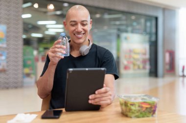 Normal ortamlarda dijital tablet bilgisayar ve salata kullanan sağlıklı yetişkin bir adam. Gıda mahkemelerinde teknoloji, beslenme ve modern yaşam tarzı arasındaki dengeyi vurgular.
