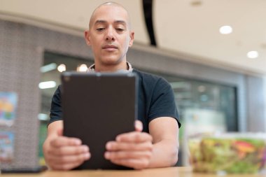 Normal ortamlarda dijital tablet bilgisayar ve salata kullanan sağlıklı yetişkin bir adam. Gıda mahkemelerinde teknoloji, beslenme ve modern yaşam tarzı arasındaki dengeyi vurgular.