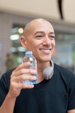 Siyah tişörtlü kel adam alışveriş merkezinde su şişesiyle su içiyor. Kamu alanındaki sağlık, kişisel bakım ve susuzluk alışkanlıklarını vurgulamak. Temiz, doğal ve asgari yaşam tarzı ifadesi.