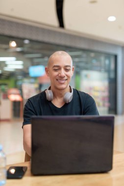 Siyah tişörtlü kel adam alışveriş merkezinde tek başına dizüstü bilgisayar kullanıyor. Toplum içinde odaklanmış dijital çalışma ve üretkenlik teması. Gerçek yaşam tarzı anı, tarafsız ve modern..