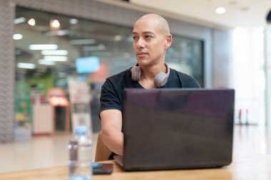 Siyah tişörtlü kel adam alışveriş merkezinde tek başına dizüstü bilgisayar kullanıyor. Toplum içinde odaklanmış dijital çalışma ve üretkenlik teması. Gerçek yaşam tarzı anı, tarafsız ve modern..