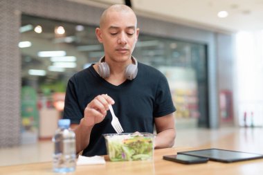 Siyah tişörtlü, kel ve yetişkin bir adam alışveriş merkezinde salata yiyor. Sağlıklı yaşam tarzı, temiz yemek ve gerçek hayatta beslenme. Doğal gün ışığı ve otantik duygu.