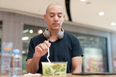 Siyah tişörtlü, kel ve yetişkin bir adam alışveriş merkezinde salata yiyor. Sağlıklı yaşam tarzı, temiz yemek ve gerçek hayatta beslenme. Doğal gün ışığı ve otantik duygu.