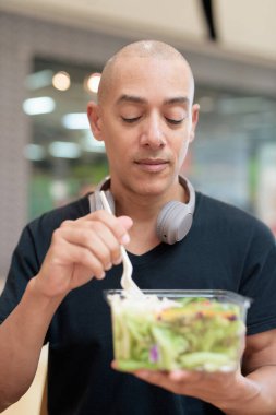 Siyah tişörtlü, kel ve yetişkin bir adam alışveriş merkezinde salata yiyor. Sağlıklı yaşam tarzı, temiz yemek ve gerçek hayatta beslenme. Doğal gün ışığı ve otantik duygu.