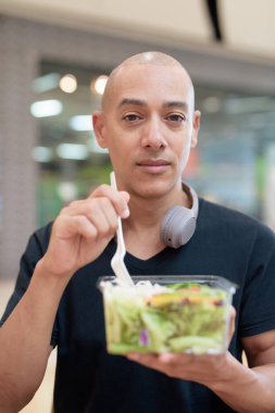 Siyah tişörtlü, kel ve yetişkin bir adam alışveriş merkezinde salata yiyor. Sağlıklı yaşam tarzı, temiz yemek ve gerçek hayatta beslenme. Doğal gün ışığı ve otantik duygu.