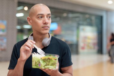 Siyah tişörtlü, kel ve yetişkin bir adam alışveriş merkezinde salata yiyor. Sağlıklı yaşam tarzı, temiz yemek ve gerçek hayatta beslenme. Doğal gün ışığı ve otantik duygu.