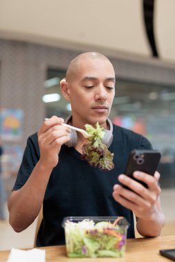 Siyah tişörtlü, kel ve yetişkin bir adam alışveriş merkezinde salata yiyor. Sağlıklı yaşam tarzı, temiz yemek ve gerçek hayatta beslenme. Doğal gün ışığı ve otantik duygu.