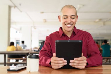 Burgundy renkli tişört giyen kendine güvenen bir adam devlet üniversitesinde ya da iş alanında kahve masasında tablet bilgisayar kullanıyor. Masadaki kahve fincanı sıradan bir hayat tarzı katıyor..
