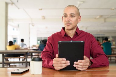 Burgundy renkli tişört giyen kendine güvenen bir adam devlet üniversitesinde ya da iş alanında kahve masasında tablet bilgisayar kullanıyor. Masadaki kahve fincanı sıradan bir hayat tarzı katıyor..
