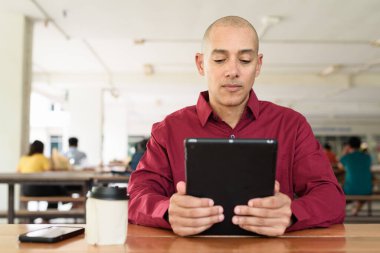 Burgundy renkli tişört giyen kendine güvenen bir adam devlet üniversitesinde ya da iş alanında kahve masasında tablet bilgisayar kullanıyor. Masadaki kahve fincanı sıradan bir hayat tarzı katıyor..