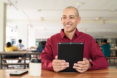 Burgundy renkli tişört giyen kendine güvenen bir adam devlet üniversitesinde ya da iş alanında kahve masasında tablet bilgisayar kullanıyor. Masadaki kahve fincanı sıradan bir hayat tarzı katıyor..