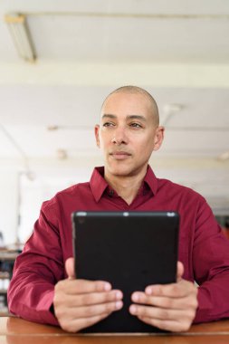 Burgundy renkli tişört giyen kendine güvenen bir adam devlet üniversitesinde ya da iş alanında kahve masasında tablet bilgisayar kullanıyor. Masadaki kahve fincanı sıradan bir hayat tarzı katıyor..