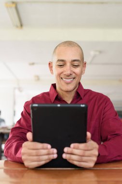 Burgundy renkli tişört giyen kendine güvenen bir adam devlet üniversitesinde ya da iş alanında kahve masasında tablet bilgisayar kullanıyor. Masadaki kahve fincanı sıradan bir hayat tarzı katıyor..